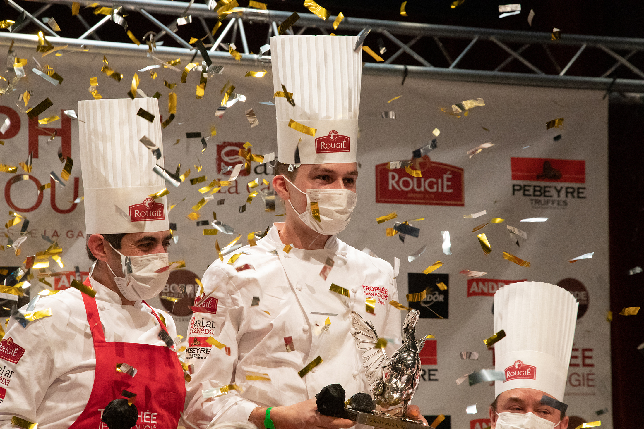 L'image montre un jeune chef cuisinier, vainqueur du prestigieux trophée Jean Rougié, prix qui lui est remis sous une pluie de paillettes dorées.