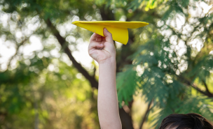 Un avion en papier jaune au centre le l’image lancé par un enfant dans une foret.
