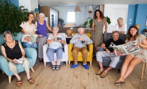 Photo de groupe d'un habitat de Domani. Certains retraités sont installés dans des fauteuils et lisent le journal, d'autres sont debout derrière eux avec les médiatrices.