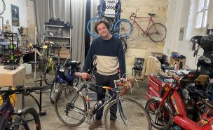 Léonard prend la pose au centre de son garage en s'appuyant sur son vélo personnel, un Peugeot. Il est entouré par de nombreux outils et d'autres engins à deux roues.