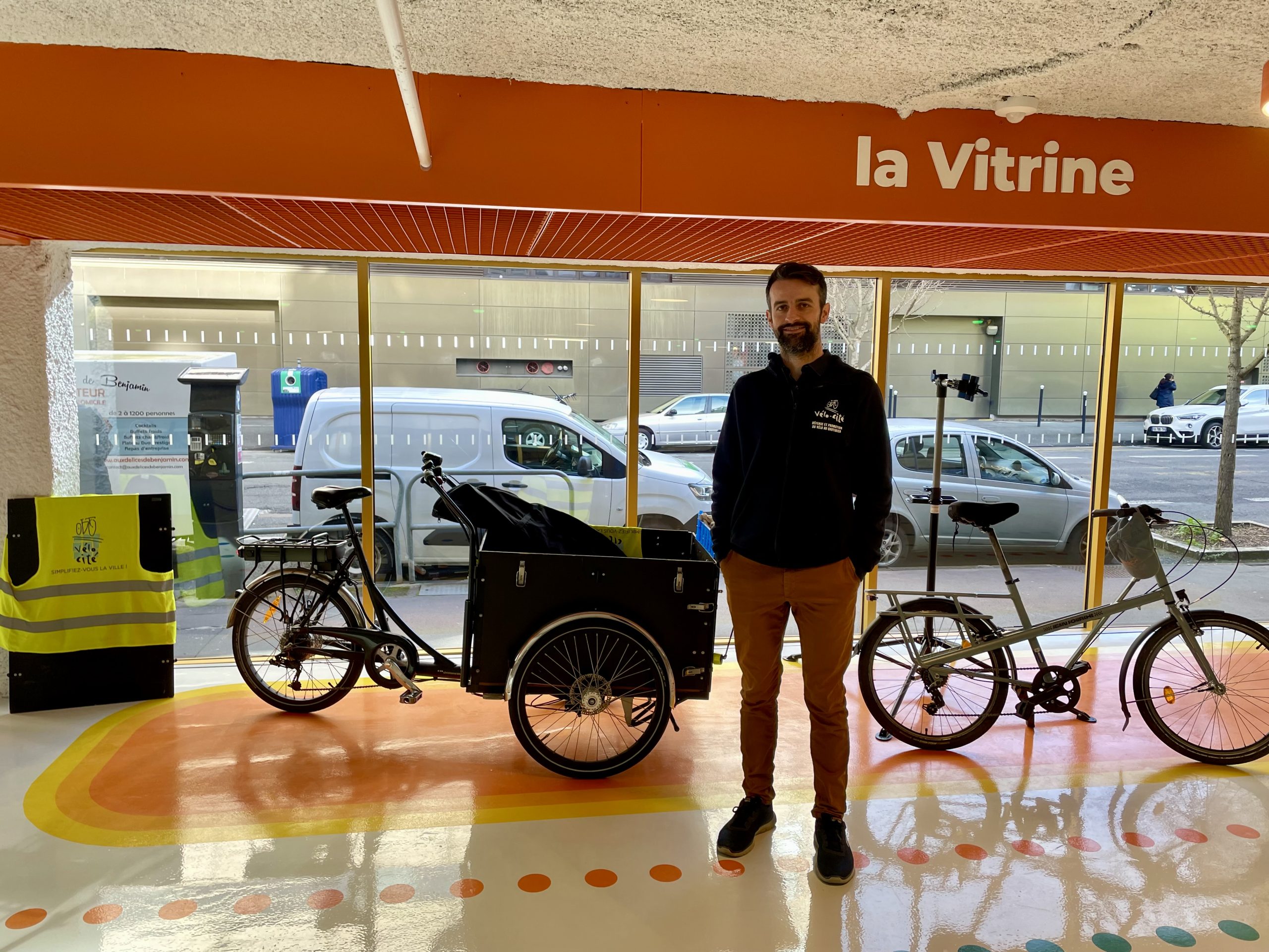 Benoît Gilliot, coordinateur de Vélo-Cité, debout devant la baie vitrée du parking. Il pose au niveau de l'espace "showroom" entouré par un vélo-cargo et une bicyclette classique.