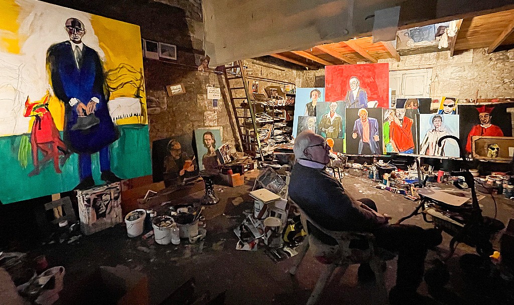 Un homme dans une atelier de peinture entouré de toiles peintes, de pots de peinture, de papier et image en tout genre.