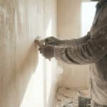Person in gloves sanding a light plastered wall; rough and smooth sections visible with dust in bright light.