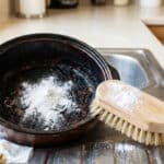 A heavily burnt saucepan in a clean kitchen sink, covered in white cleaning powder. A wooden scrubbing brush lies nearby, dusted with powder.
