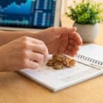 Hands placing euro coins and banknote on an "Avril 2026" calendar. Blurred tablet with financial data and plant in background.