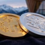 Detailed gold and silver Olympic medals on dark velvet. Gold has snowflakes and arches; silver shows skiers, gondola. Blurred mountains behind.