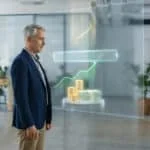 Un homme mature regarde un écran holographique affichant des graphiques de croissance financière, des pièces et des billets, en bureau.