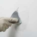 A gloved hand expertly smooths white joint compound on a wall repair with a silver trowel, aiming for an invisible finish.