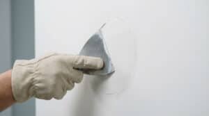 A gloved hand expertly smooths white joint compound on a wall repair with a silver trowel, aiming for an invisible finish.
