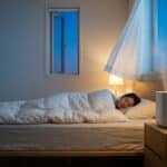 A woman sleeps peacefully in a modern bedroom at night. An open window, swaying sheer curtain, and a humidifier emitting mist suggest fresh, clean air.