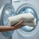 Main tenant des serviettes blanches moelleuses devant une machine à laver ouverte, tambour éclairé. Linge propre.