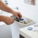 Person's hands replacing a blue rubber washer in a toilet tank with pliers, showing a practical home repair in a clean bathroom.