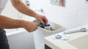 Person's hands replacing a blue rubber washer in a toilet tank with pliers, showing a practical home repair in a clean bathroom.