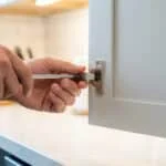 Gros plan sur des mains ajustant précisément une charnière de porte d'armoire de cuisine blanche avec un tournevis.