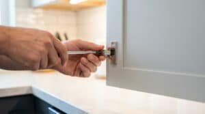 Gros plan sur des mains ajustant précisément une charnière de porte d'armoire de cuisine blanche avec un tournevis.