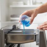 Une main verse du détergent bleu d'un gobelet doseur dans le bac d'une machine à laver grise, soulignant le dosage correct.