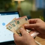 Mains tenant des billets et pièces en euros devant une tablette affichant le calendrier d'avril 2026.