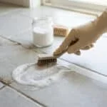 Main gantée frottant un joint de carrelage noirci avec une brosse, à côté d'une solution moussante et un pot de poudre blanche.