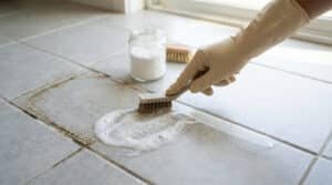 Main gantée frottant un joint de carrelage noirci avec une brosse, à côté d'une solution moussante et un pot de poudre blanche.