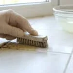 Main gantée frottant des joints de carrelage encrassés avec une brosse. Un bol de solution nettoyante est à côté.