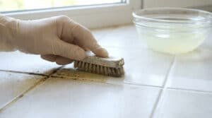 Main gantée frottant des joints de carrelage encrassés avec une brosse. Un bol de solution nettoyante est à côté.