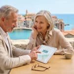 Un couple senior discute de sa retraite 2026 en consultant une tablette sur une terrasse ensoleillée, mer et village en arrière-plan.