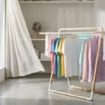 Un séchoir à linge moderne en bois et métal, chargé de vêtements colorés, près d'une fenêtre ouverte avec un rideau flottant et un ventilateur.