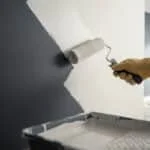 Main gantée appliquant un apprêt teinté clair avec un rouleau sur un mur gris foncé, l'éclaircissant progressivement.