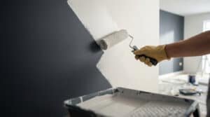 Main gantée appliquant un apprêt teinté clair avec un rouleau sur un mur gris foncé, l'éclaircissant progressivement.