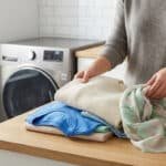 Mains pliant des vêtements propres (pull, t-shirt, foulard) sur un comptoir en bois, avec une machine à laver en arrière-plan.