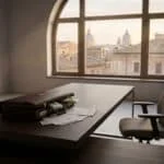 Une mallette ouverte pleine de billets sur un bureau moderne. Des documents et une chaise sont visibles, avec vue urbaine par la fenêtre.