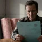 Un homme préoccupé examine un document bleu, une main gantée près de lui. Des rouleaux d'isolant rose sont en arrière-plan.