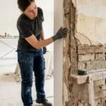 Femme en gants de travail inspectant un mur ancien dénudé, révélant la structure interne (briques, bois) lors de travaux.