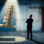 Homme en costume face à une pile chancelante de monnaie sur un échafaudage, symbolisant la dette publique et son instabilité.