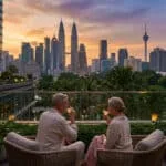 Couple de retraités profitant d'un verre sur une terrasse luxueuse à Kuala Lumpur, avec les tours Petronas et la tour KL au coucher du soleil.