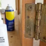 Un tube de graisse au lithium blanche devant une charnière de porte en laiton avec un aérosol WD-40 en arrière-plan.