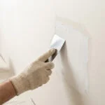 Une main gantée utilisant une spatule pour étaler de l'enduit sur un mur en plaques de plâtre.