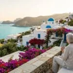 Une personne âgée assise sur une terrasse fleurie surplombant un village grec blanc avec des églises à dôme bleu.