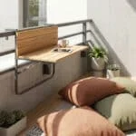 Une table pliante en bois fixée à la rambarde d'un balcon avec des coussins au sol et des plantes vertes.