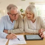 Un couple de seniors examine attentivement des documents financiers et une tablette sur une table dans un salon lumineux.