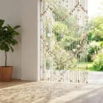 Un rideau de perles en bois et nacre suspendu devant une porte-fenêtre ouverte donnant sur un jardin ensoleillé.
