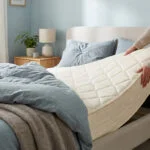 Une personne soulève et fait pivoter un matelas blanc sur un lit dans une chambre lumineuse et moderne.
