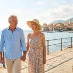 Un couple de seniors souriants se promène main dans la main sur une jetée ensoleillée près d'un village méditerranéen.