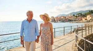Un couple de seniors souriants se promène main dans la main sur une jetée ensoleillée près d'un village méditerranéen.