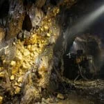 Une paroi rocheuse dans une mine sombre éclairée par des projecteurs, révélant des veines d'or brillantes incrustées.