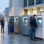 Des personnes utilisent des distributeurs de billets alignés sur une place urbaine moderne.