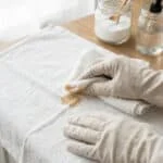 Une personne avec des gants nettoie une tache sur un tissu blanc avec un chiffon, entourée de produits ménagers naturels.