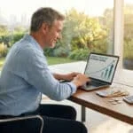 Un homme souriant consulte des projections d'épargne retraite sur un ordinateur portable devant une fenêtre ensoleillée.