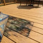 Une main soulève un tapis extérieur révélant du bois pourri et de la mousse sur une terrasse en bois.