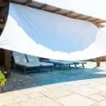 Un drap blanc tendu sous une pergola en bois pour créer de l'ombre sur une terrasse en pierre avec des transats.